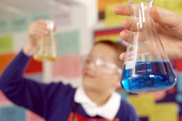 A hand holds a glass beaker of blue liquid, boy in googles looks on 